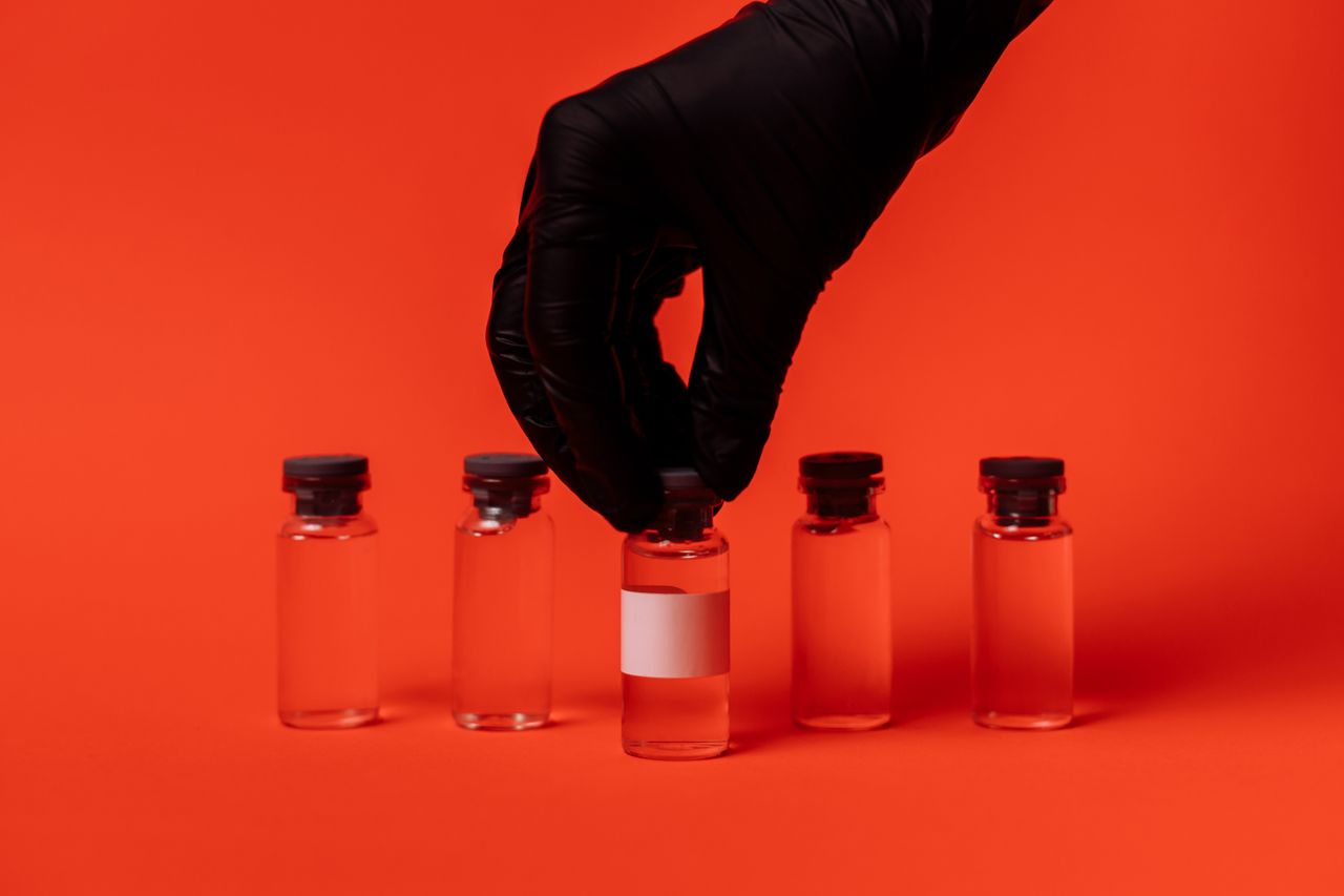 Person wearing black glove grabbing a vaccine vial against a bright red backdrop.