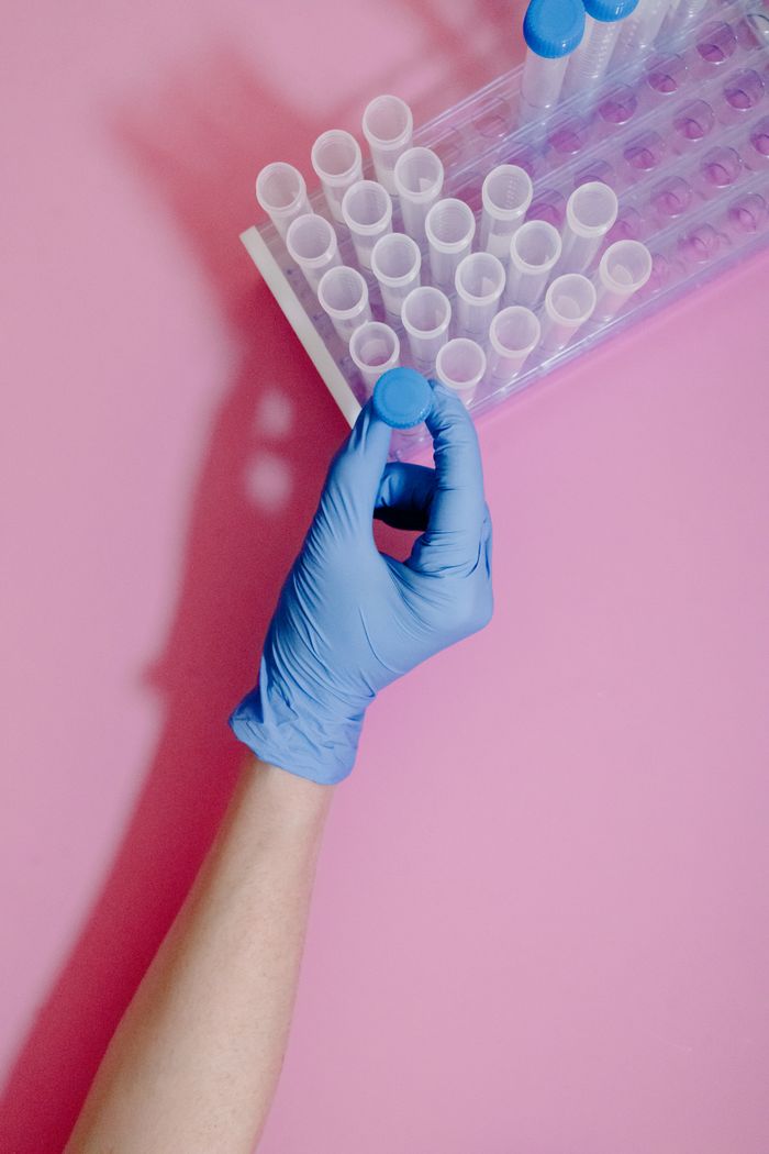 A gloved hand holding a blue-capped test tube on a pink background.